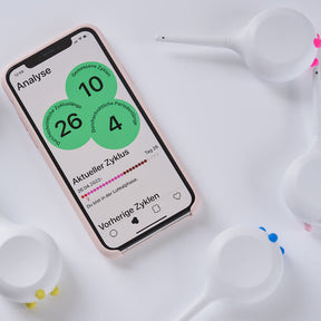 Mobiltelefon zu sehen, das die Oberfläche der Teena App zeigt. Es ist umgeben von Teena Period-Trackern, die weiß sind und Füßen in verschiedenen Farben haben.